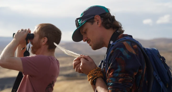 Two adults outdoors, one examining a feather and the other looking through binoculars